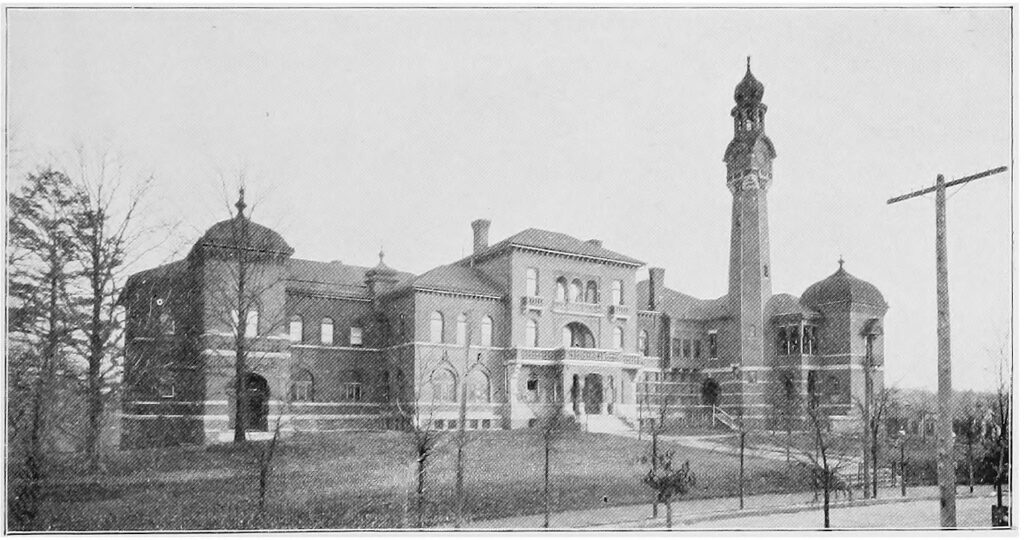 Black and white photograph of a large building compound with asymmetrical  towers with rounded roofs on both the left and right wings.