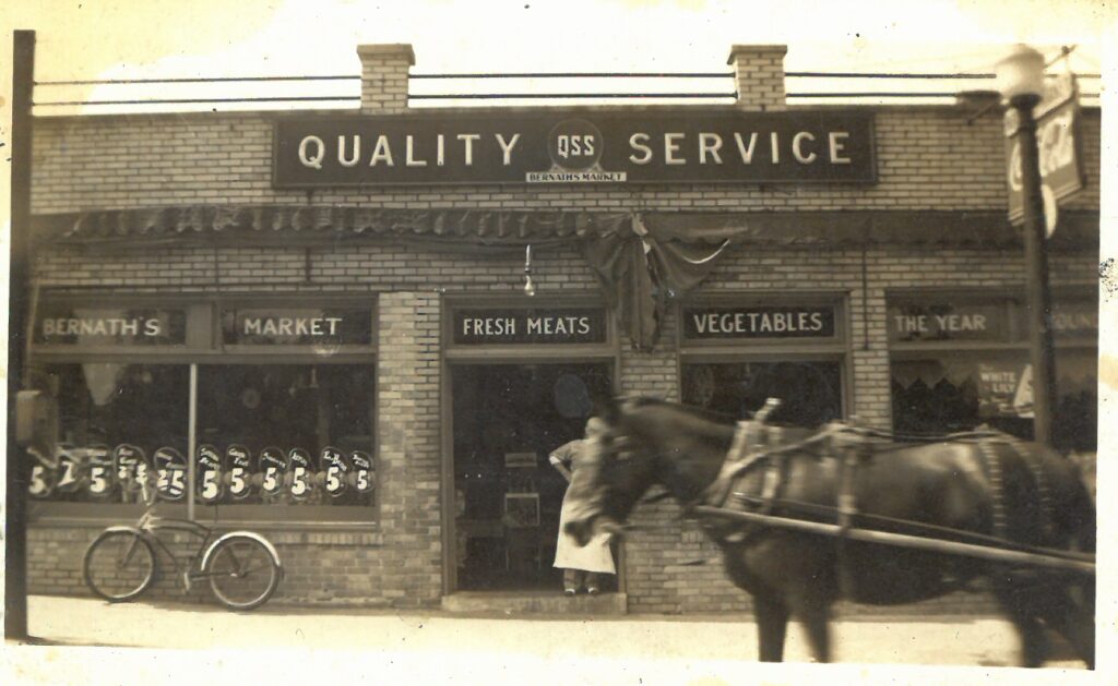 Sepia-tone photograph showing a brick store facade with a sign that reads "Quality Service" over window displays that read "Bernath's Market Fresh Meats Vegetables." There is a bicycle parked beneath one window and a horse in the foreground.