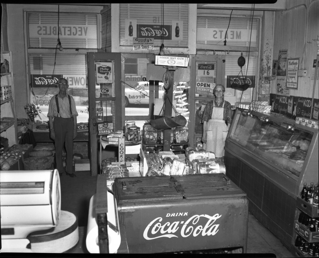 Black and white photograph of the interior of a grocery store showing two older male clerks beside product cases