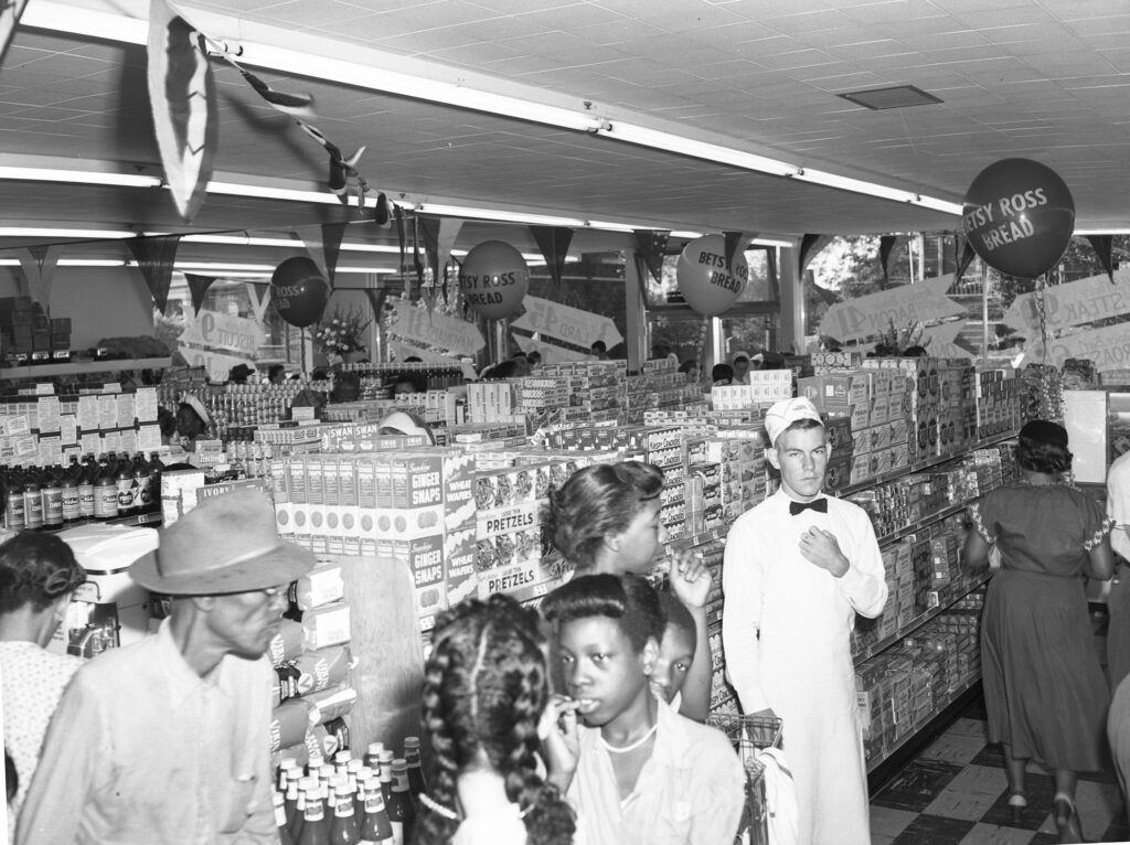 Black and white photograph of the interior of a grocery store showing a uniformed clerk beside a group of customers in the foreground and aisles of products in the background. The store is decorated with balloons and bunting.