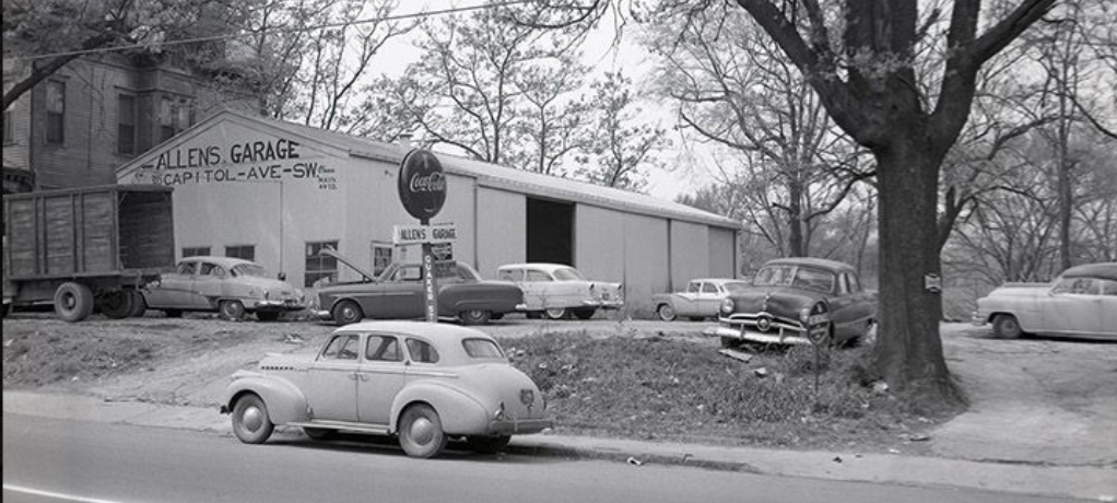 Black and white photograph of a white shed-like building surrounded by cars.