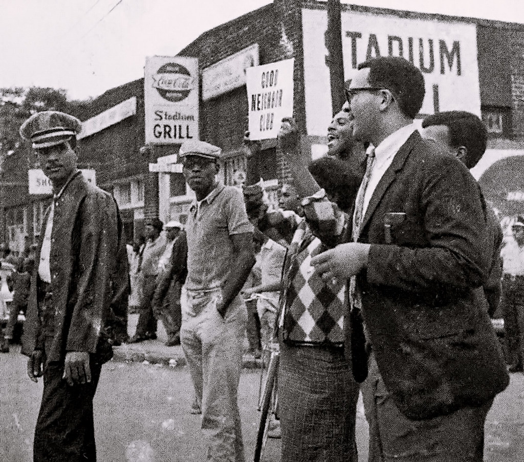 Black and white photograph showing men gathered, holding signs and shouting.