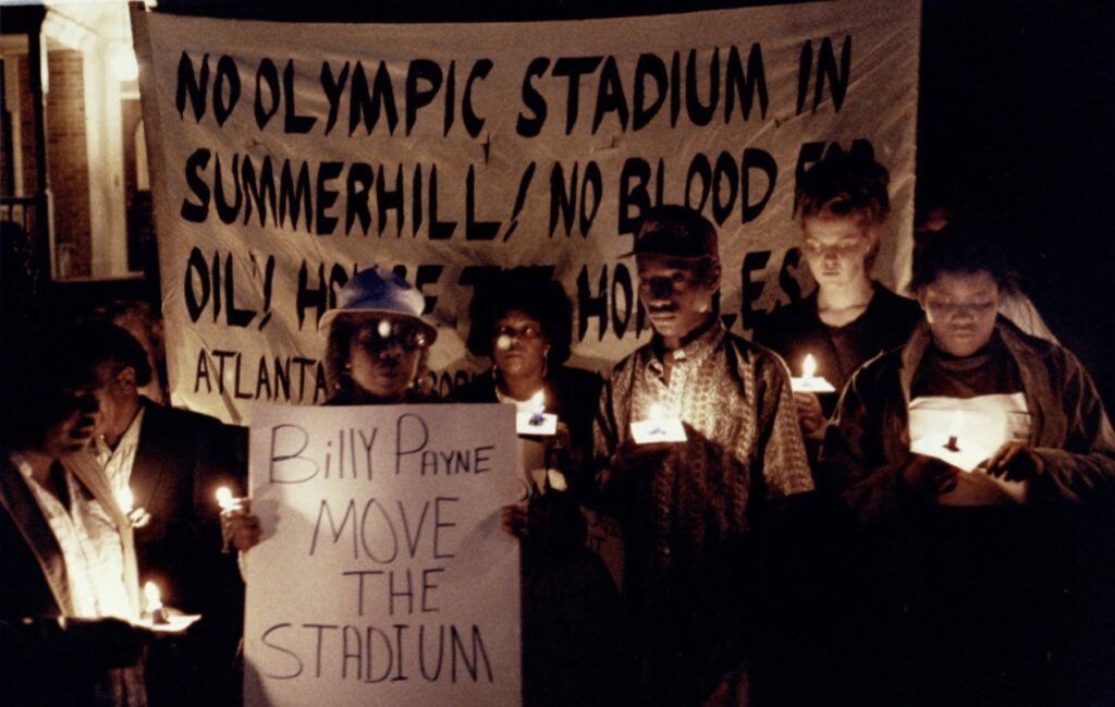 Color photograph of a group of people holding signs and candles at night. Visible behind them is a banner with words "No Olympic Stadium In Summerhill"