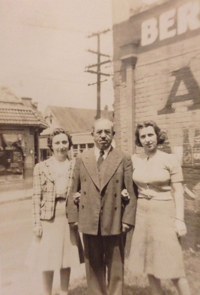 Sepia tone photograph of an older man wearing a coat and tie standing between two young women. 