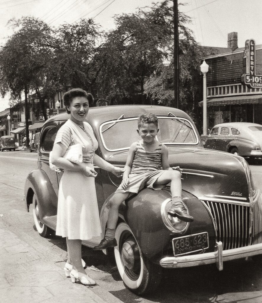 Black and white photograph of a young woman wearing a light-colored dress with a boy seated on the fender of a car. 