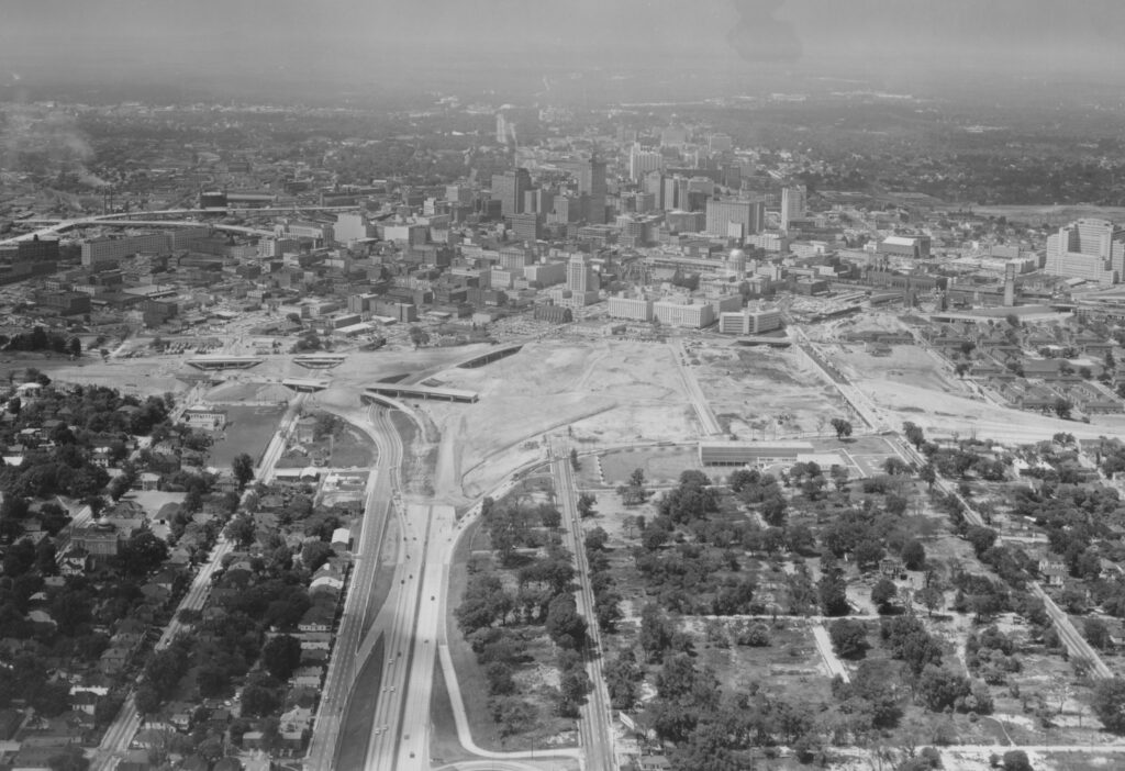 Black and white photograph showing a large amount of cleared, flat land with skyscrapers in the background and two separate areas of trees and buildings in the foreground, separated by a wide road. 