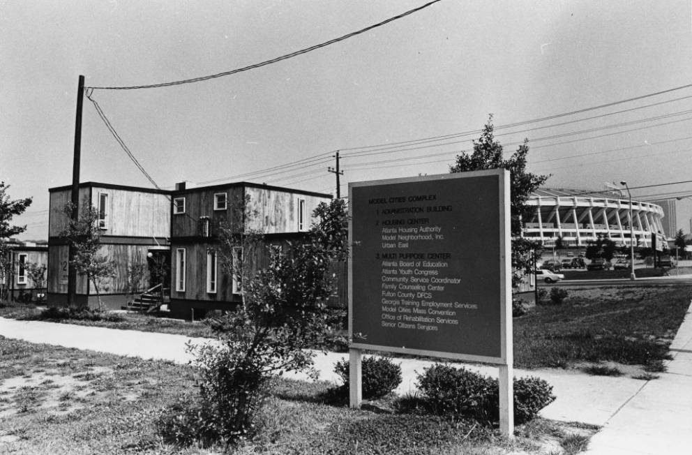 Black and white photograph showing small rectangular structures behind a large sign, with the stadium in the background.