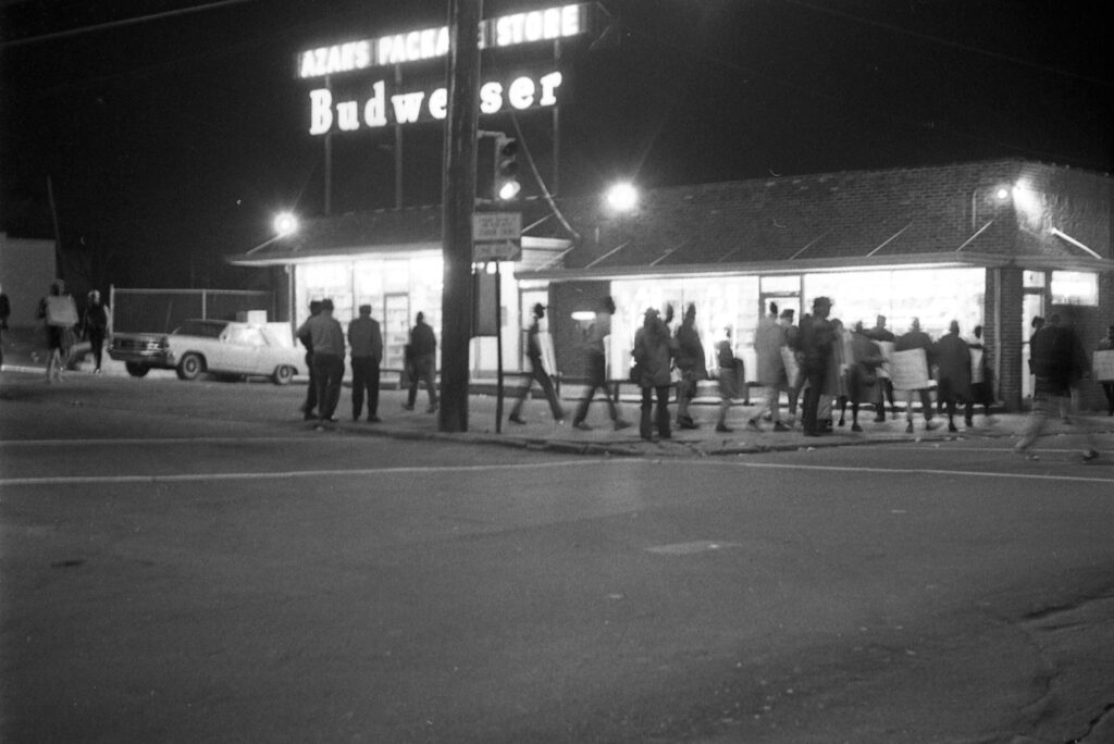Black and white photo of a group of people lined up in front of Azar's Package store at night; the store is lit up with a neon sign advertising Budweiser