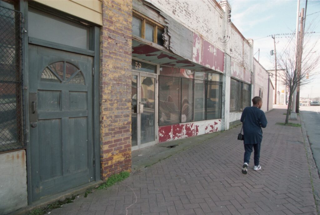 Color photograph of a person walking along a brick sidewalk in front of empty stores