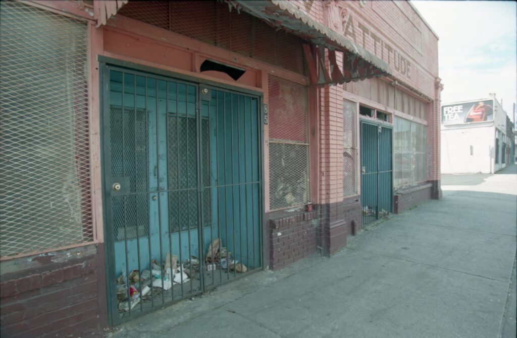 Color photo of a blue-painted storefront with bars over the entrance and trashed piled up behind the bars