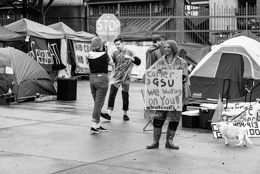 A protester holds up a sign that reads "Camer & GSU We Waiting on You!" in front of a group of tents.