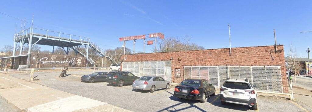 Color photograph of a brick storefront with barred windows and cars parked in front of it. A building across the way has a mural painted on the side that says "Summerhill"