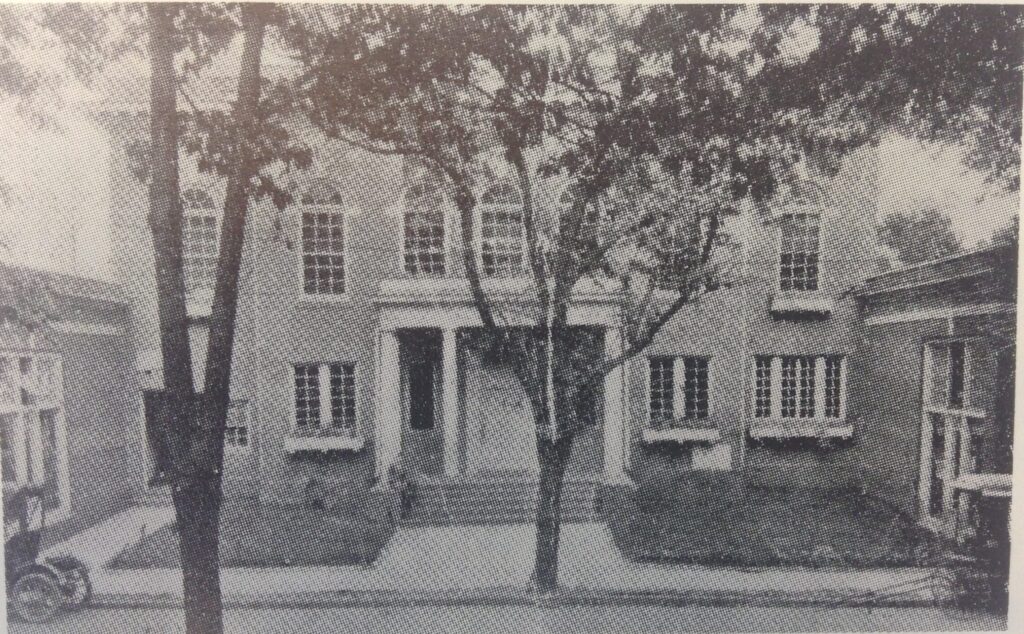 Black and white photograph of a large brick building with many arched windows on the second floor and a rectangular portico with four columns above the central door.