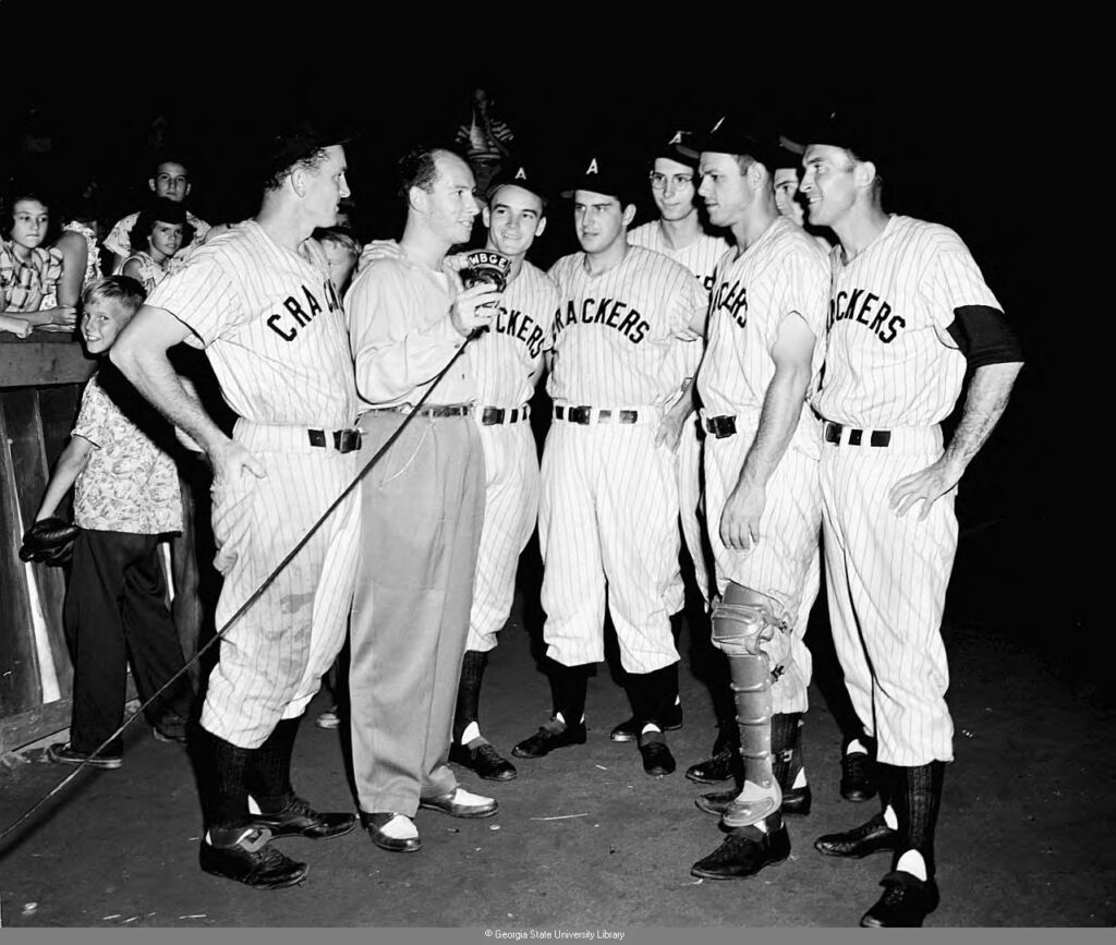 Black and white image of seven members of the Atlanta Crackers baseball team with a WSB radio reporter with children and others in the background