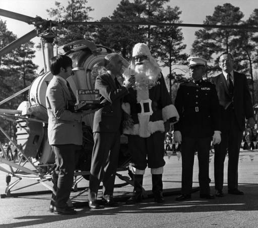 black and white image of a radio reporter and three other men with a Santa Claus with a helicopter in the background