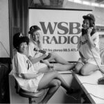 black and white image of three women in a press booth for WSB radio at an Atlanta Braves baseball game. one is pretending to be on the phone and the another is holding a WSB microphone pointed at the third