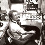 black and white of a man in a radio station booth with equipment around him, turntables, to reel-to-reel, to Fidelipac radio carts 