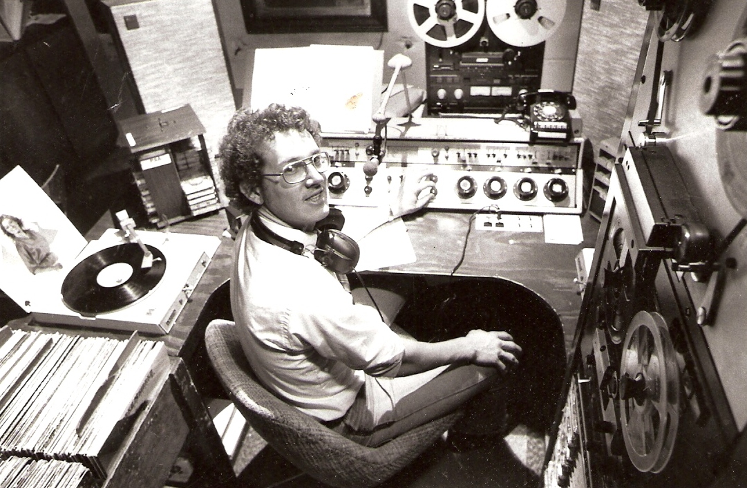 black and white of a man in a radio station booth with equipment around him, turntables, to reel-to-reel, to Fidelipac radio carts 