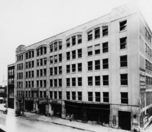 Black and white photo of a seven-story tall building with rows of square windows and store-fronts on the bottom level