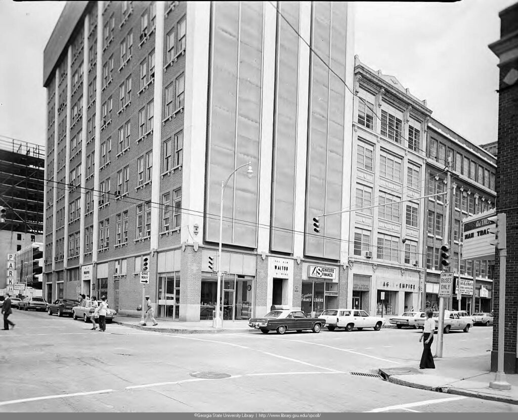 Black and white image of a street corner with a seven-story tall building on it with cars parked in front.